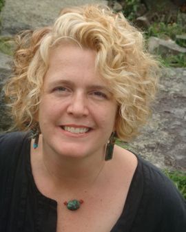 Headshot of Juanita Rockwell from when she was in her 50s. She has blond curly hair, greying at the front, fair skin and is smiling. She is wearing ling dangly earrings ad a necklace with 1 jade bead framed by two smaller red beads. She is wearing a black open-necked shirt and is against a natural background.